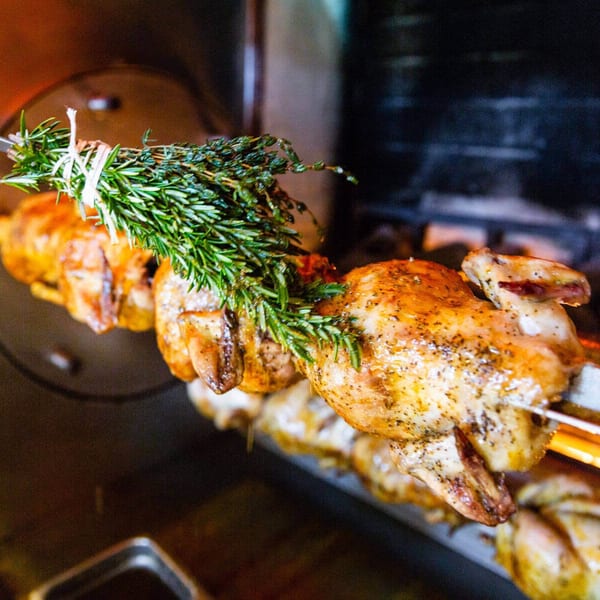 Golden-brown rotisserie chickens with fresh rosemary and thyme bundles cooking over an open flame.