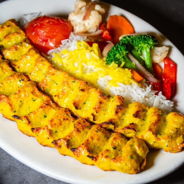 Two skewers of golden chicken kabob with white and yellow rice, roasted tomato, and mixed vegetables.