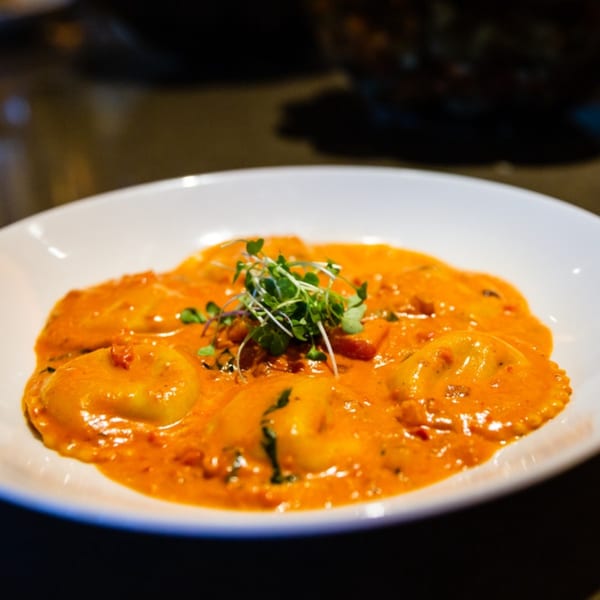 Ravioli pasta in a creamy orange sauce, garnished with fresh microgreens in a white bowl.