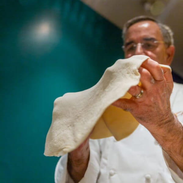 Chef stretching pizza dough by hand against a teal background at Faz Restaurants & Catering.