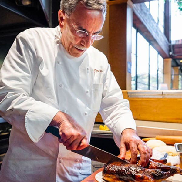 A chef in a white coat and glasses carves a roasted bird on a wooden counter at Faz Restaurants.