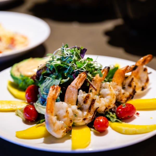 Grilled shrimp skewers with mixed greens, avocado, cherry tomatoes, and mango slices on a white plate.