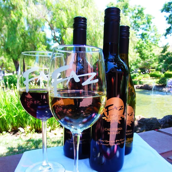 Faz Restaurant's branded wine bottles and glasses with red and white wine, outdoors by a pond.