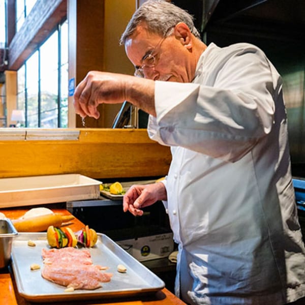A chef in a white coat sprinkles seasoning over raw chicken and vegetable skewers on a baking sheet.