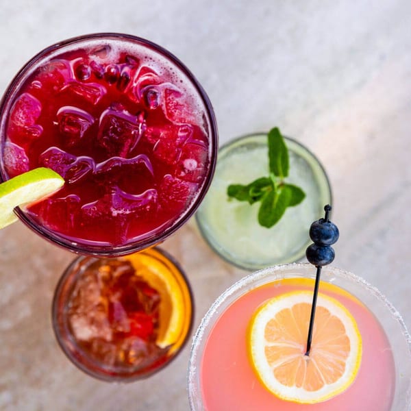 Overhead view of four colorful cocktails, including red, green, orange, and pink drinks with garnishes.