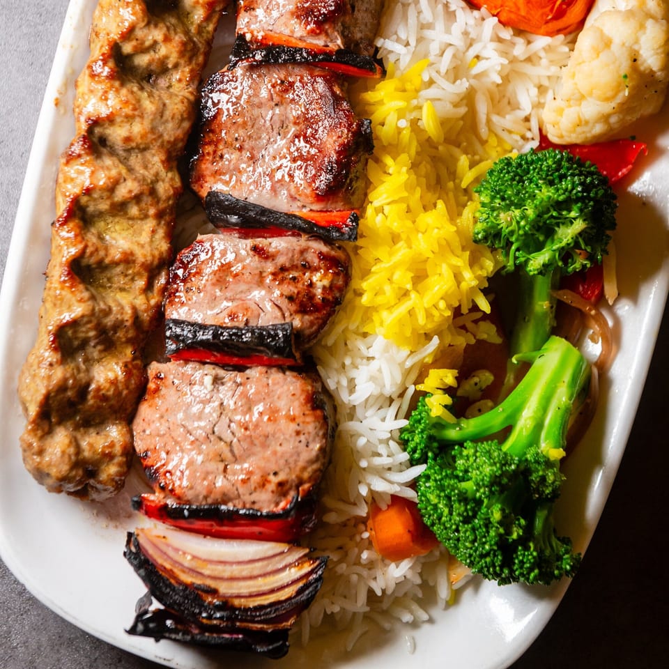 Mediterranean platter with two skewers of grilled meat, yellow and white rice, and steamed broccoli.